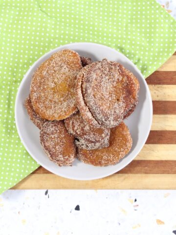 A close-up of pumpkin fritters coated in cinnamon sugar, one with a bite showing its bright orange, fluffy center, on a white plate over a green dotted napkin.