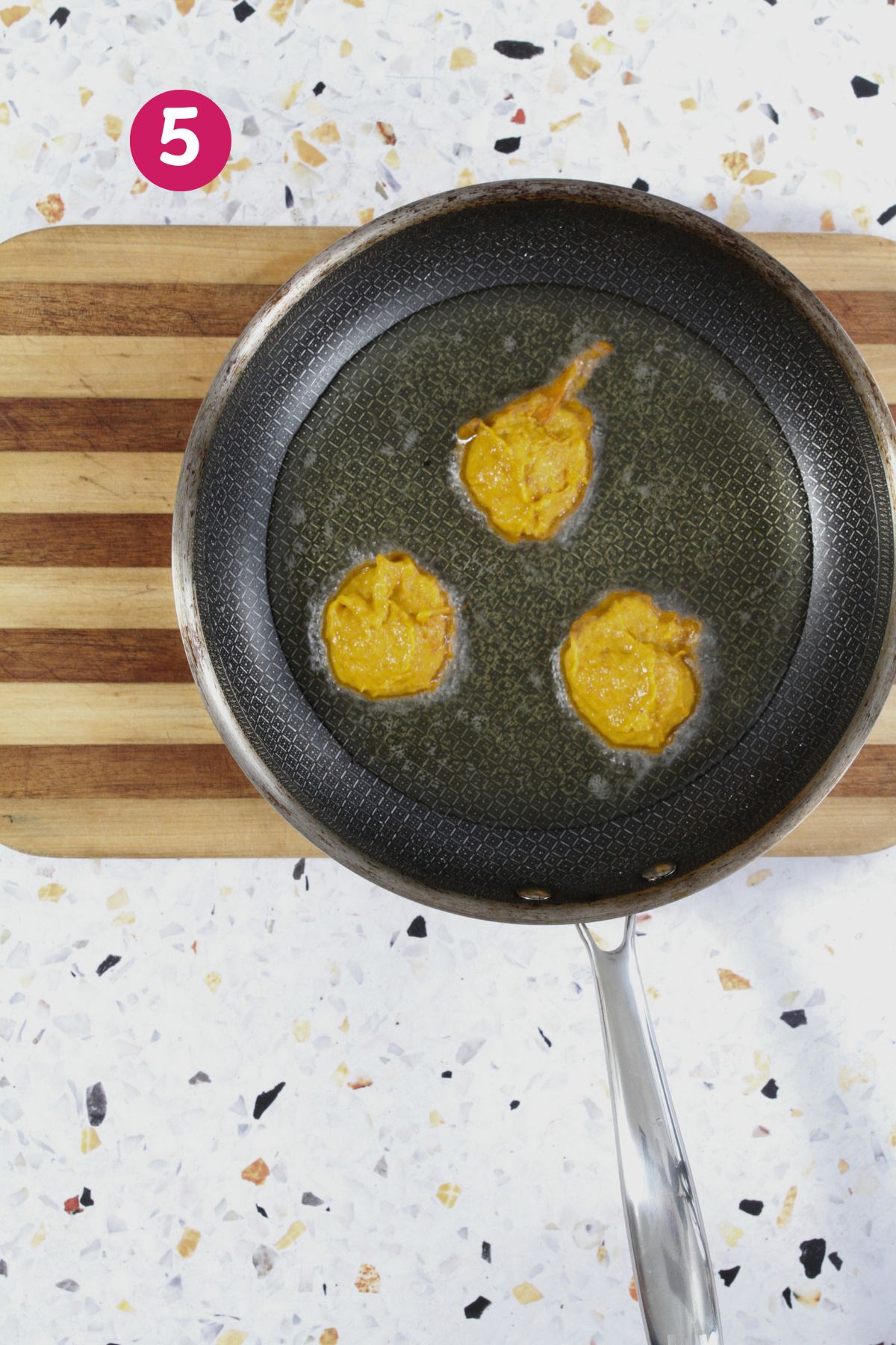 Three spoonfuls of pumpkin batter are frying in hot oil in a skillet on a wooden board, labeled with a pink