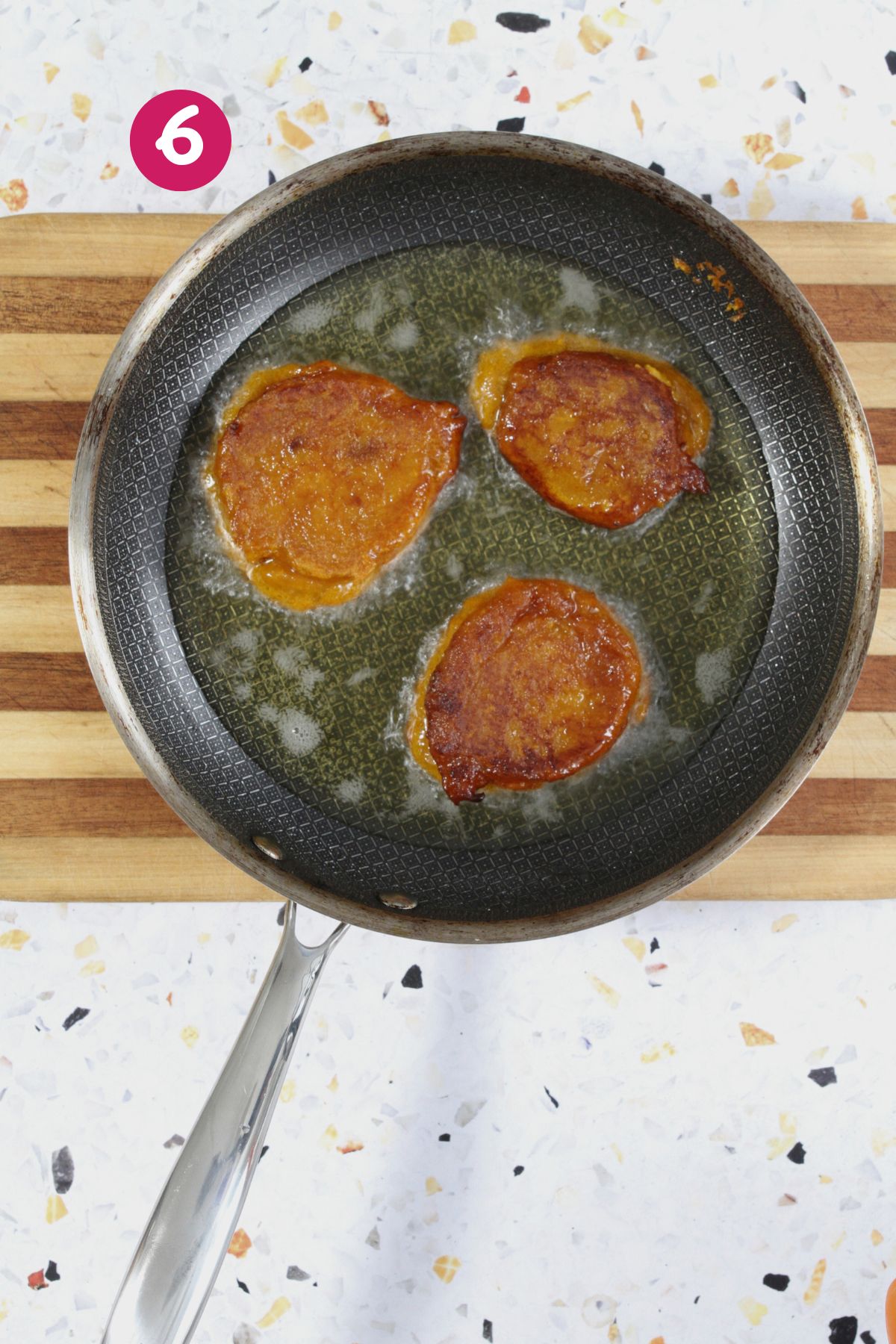 Three golden-brown fritters sizzle in a skillet of oil, showing the frying process, labeled with a pink