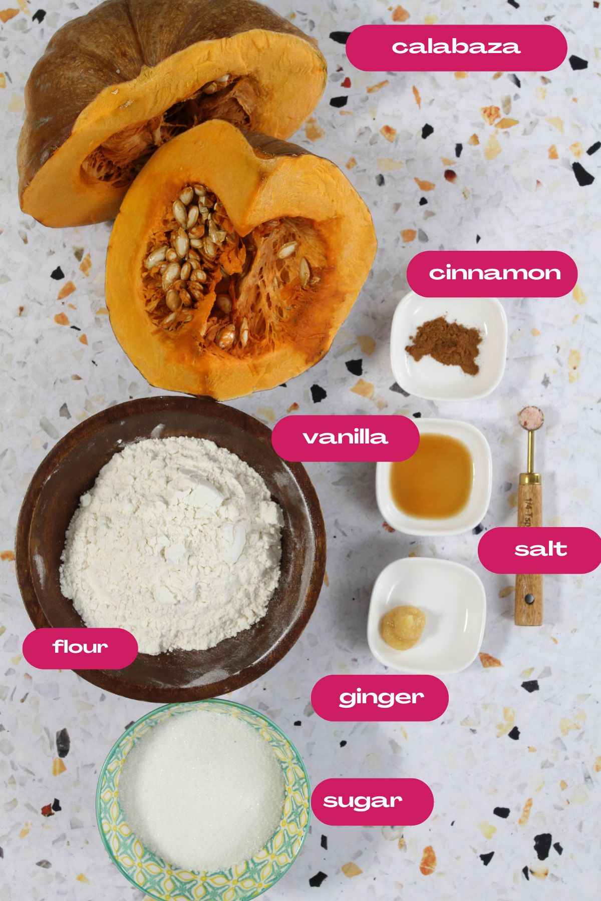 Ingredients for pumpkin fritters arranged on a light speckled surface: halved calabaza, flour, sugar, cinnamon, ginger, salt, and vanilla extract, each labeled with pink tags.