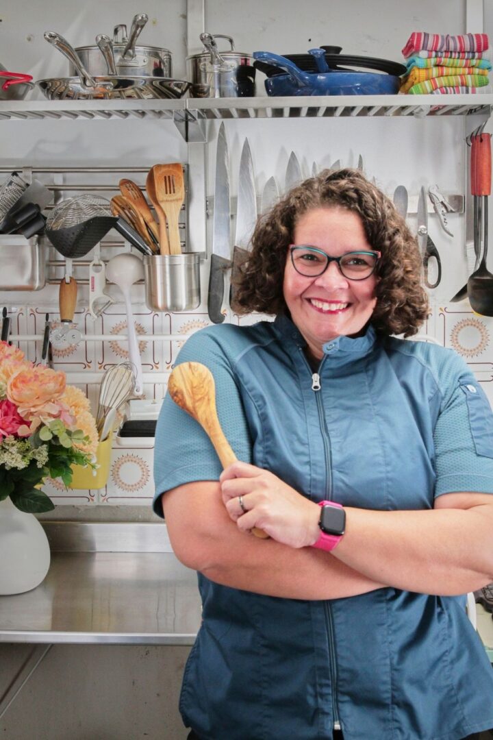 A woman with short curly hair smiles and holds a wooden spoon in a cheerful kitchen with flowers and utensils behind her.
