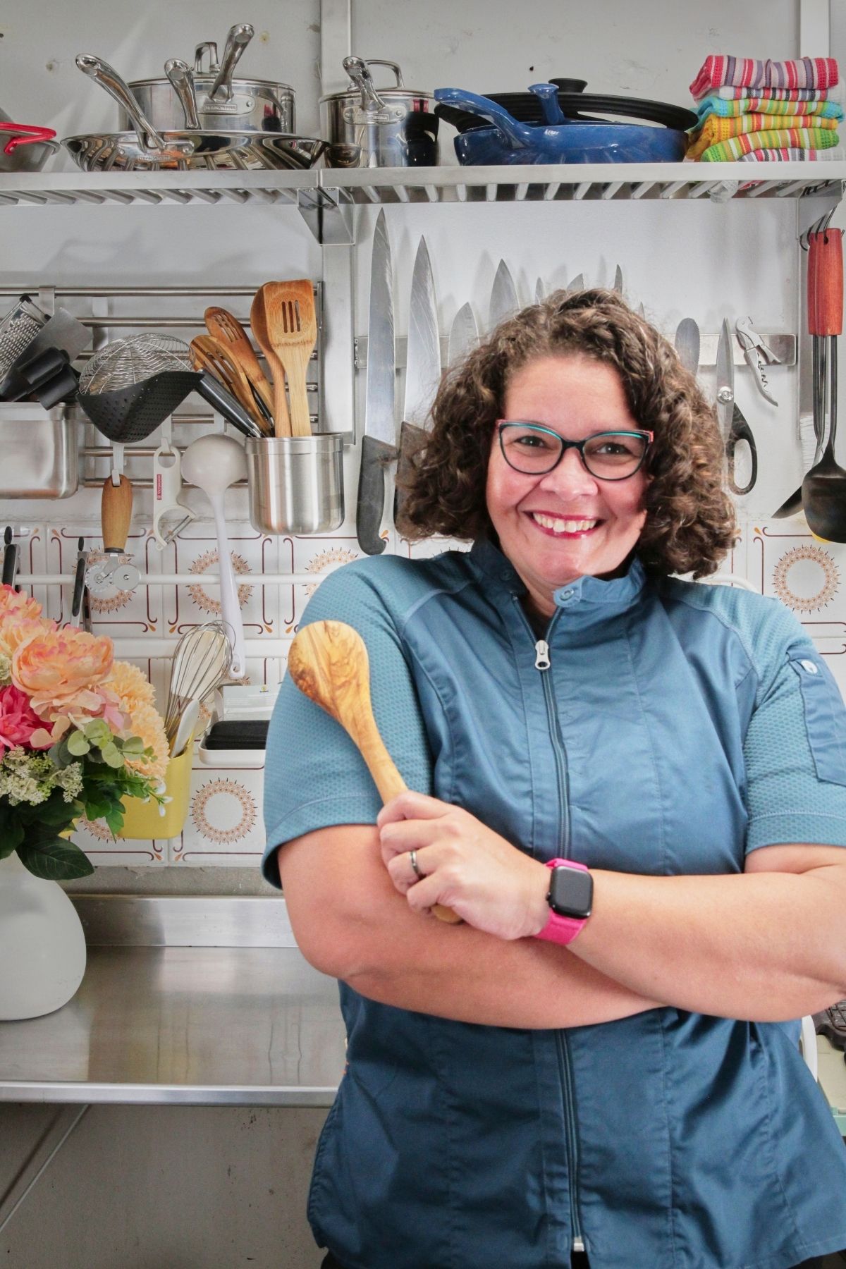 A woman with short curly hair smiles and holds a wooden spoon in a cheerful kitchen with flowers and utensils behind her.