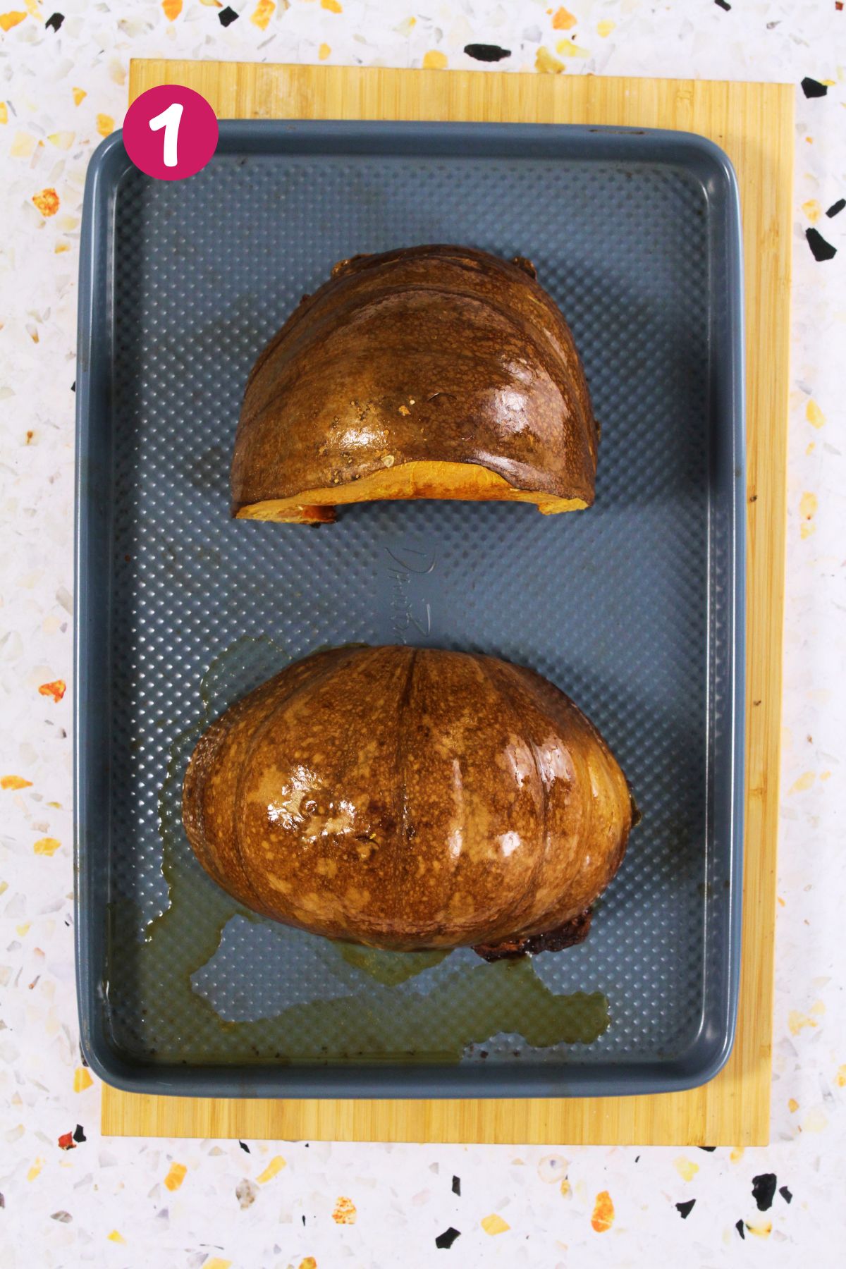Two large pieces of roasted calabaza with browned skin sit on a baking sheet lined with oil, showing the first step of preparing pumpkin fritters.