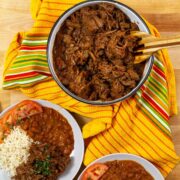 Bowl of Puerto Rican ropa vieja, shredded beef stew with sofrito and sazón, served alongside rice, beans, and fresh tomato slices.