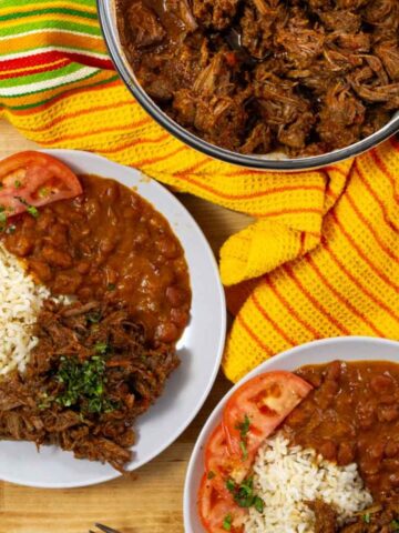 Overhead view of Puerto Rican ropa vieja (shredded beef stew) with white rice, stewed beans, and tomato slices, on a plate.