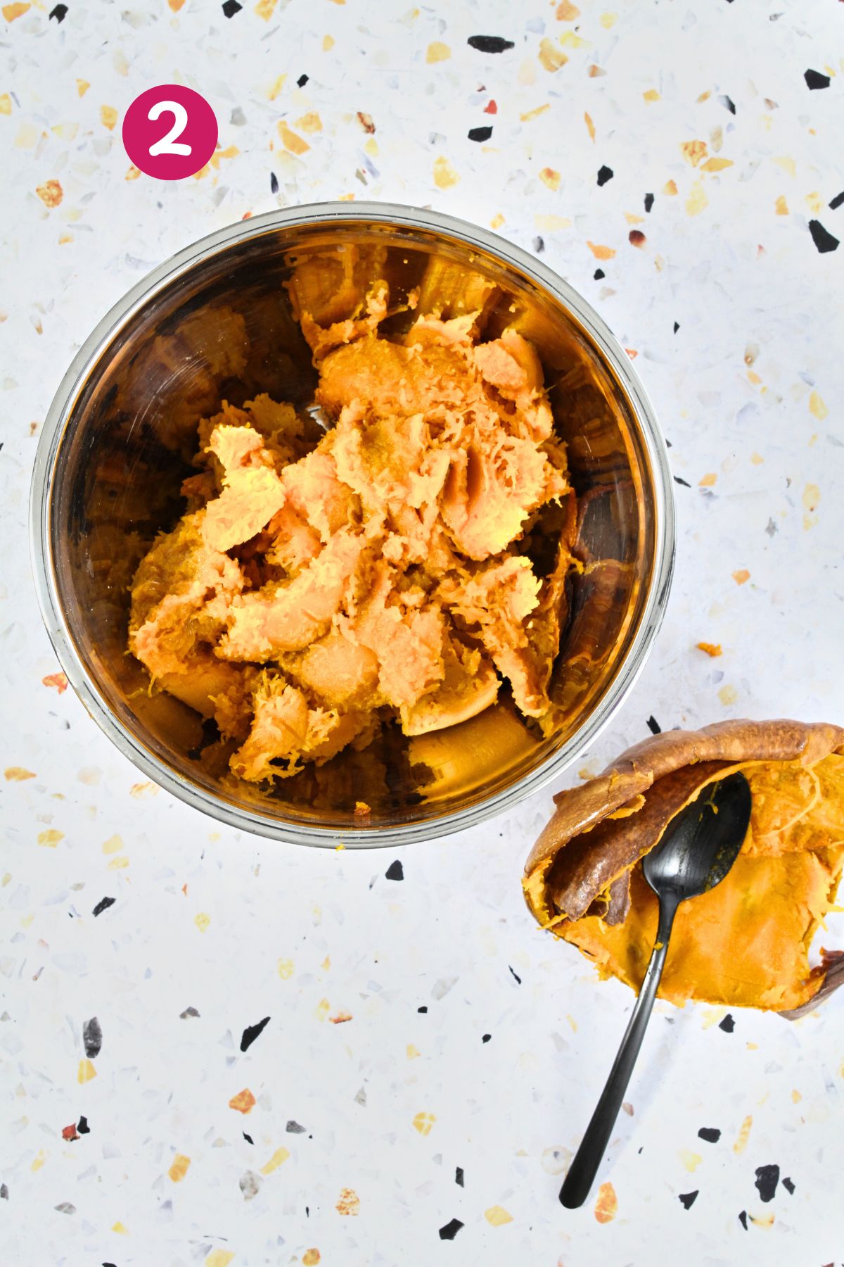 A metal bowl filled with scooped roasted pumpkin pulp sits beside the empty pumpkin skin and a black spoon, ready for mixing.