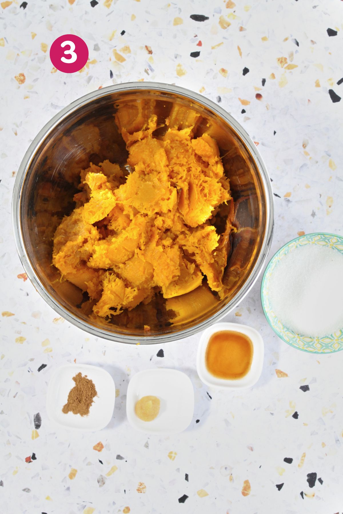 A metal bowl of mashed pumpkin is surrounded by small bowls of sugar, cinnamon, ginger, and vanilla extract, showing the seasoning step for fritter batter.