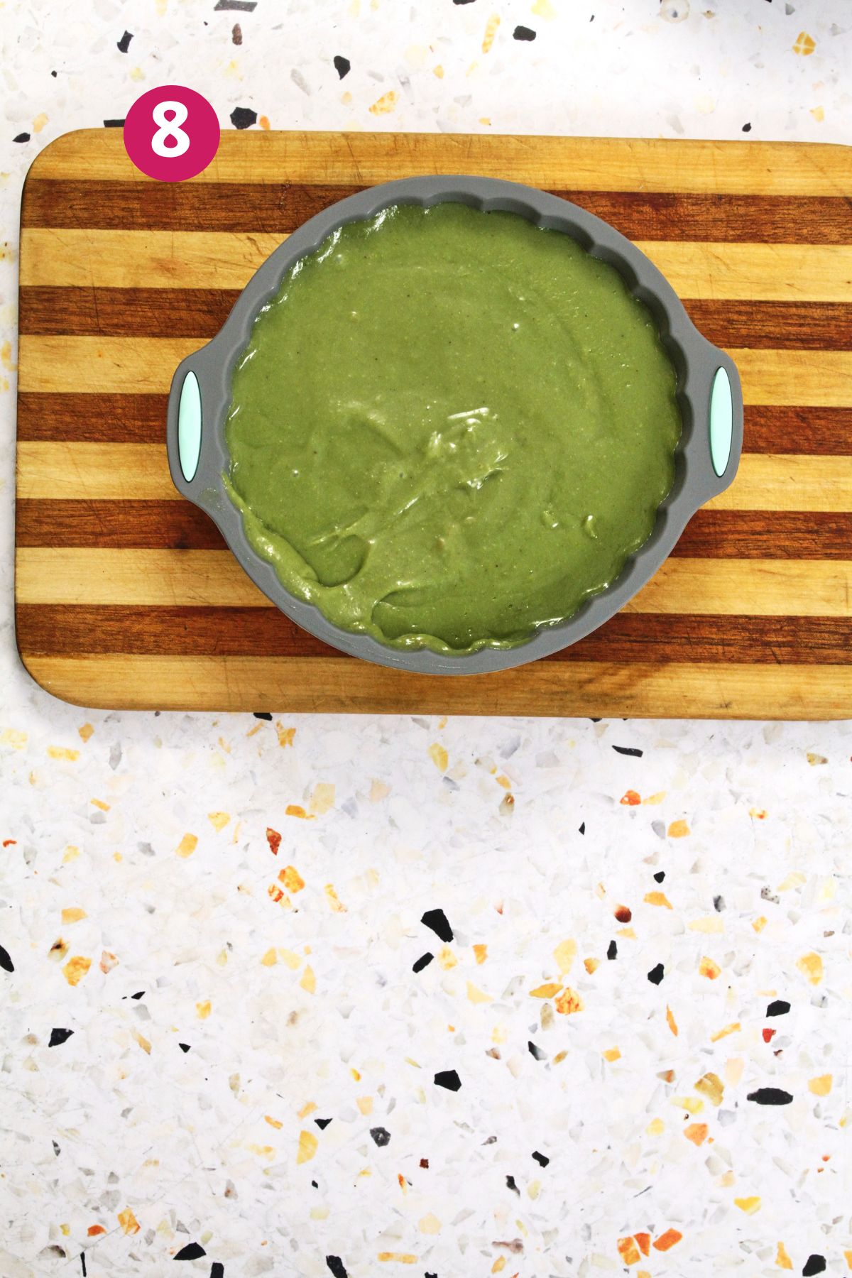 Green pistachio tembleque mixture poured into a silicone mold resting on a striped wooden cutting board.