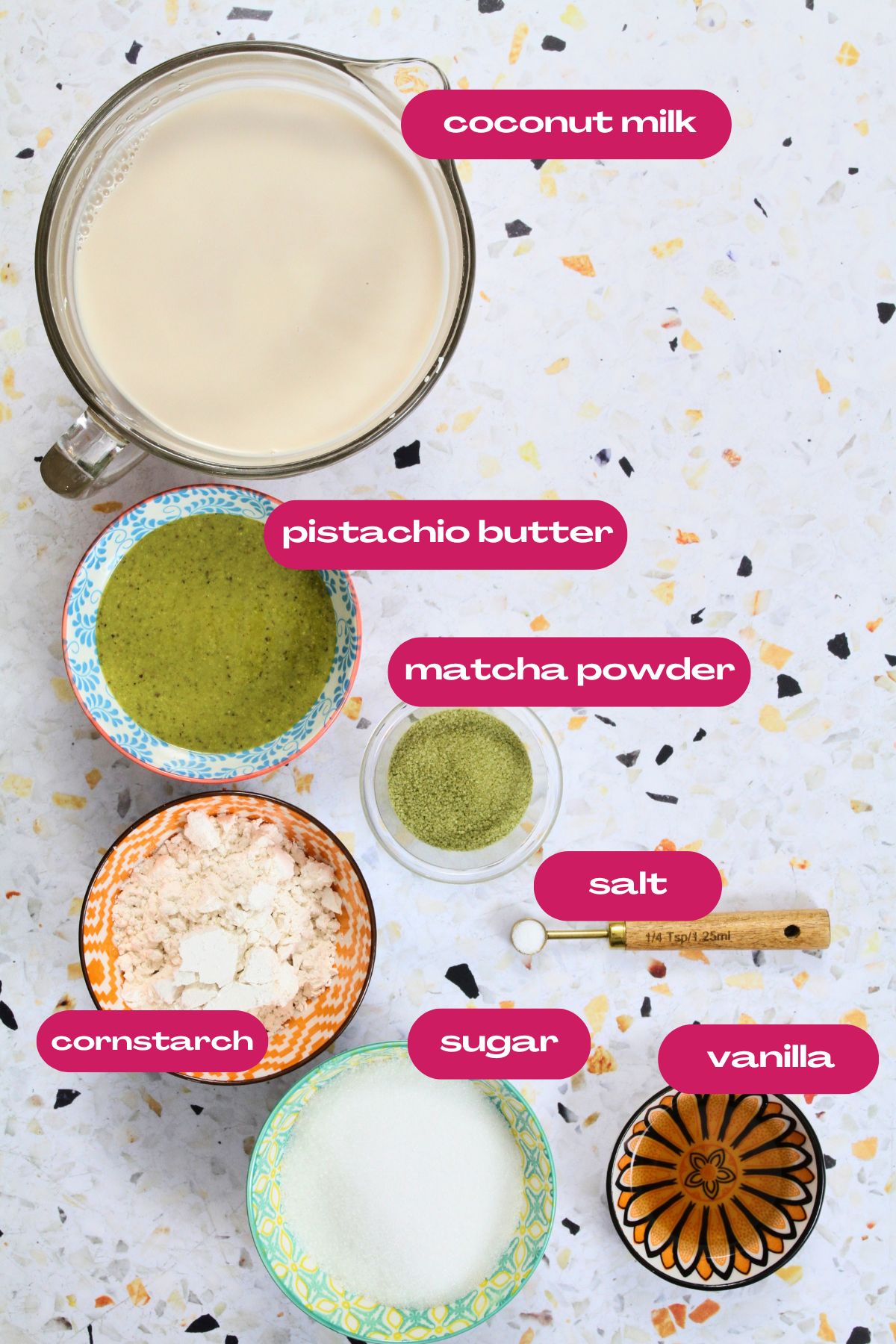 Labeled ingredients for pistachio tembleque arranged on a terrazzo surface, including coconut milk, pistachio butter, matcha powder, cornstarch, sugar, salt, and vanilla.