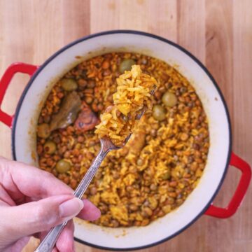 Spoon lifting arroz con gandules with rice, pigeon peas, olives, and pork from a pot.