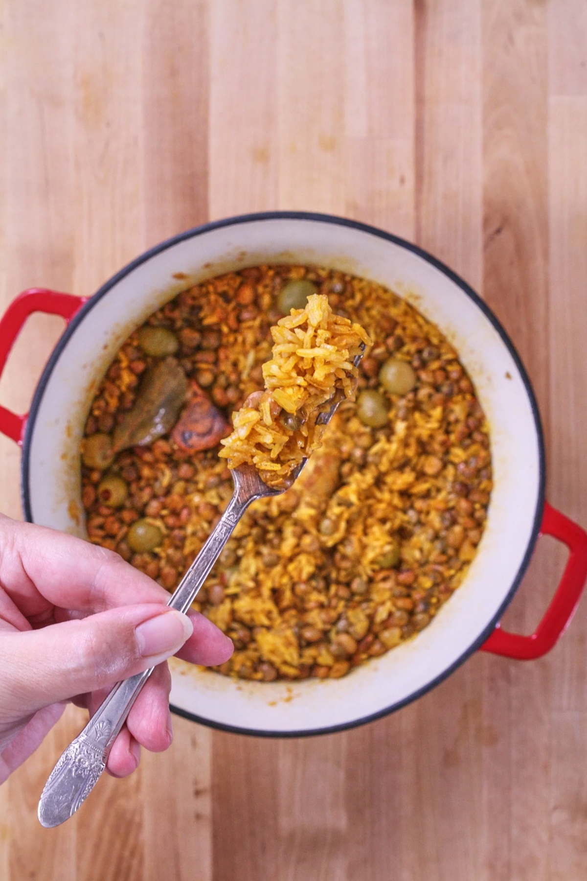 Spoon lifting arroz con gandules with rice, pigeon peas, olives, and pork from a pot.