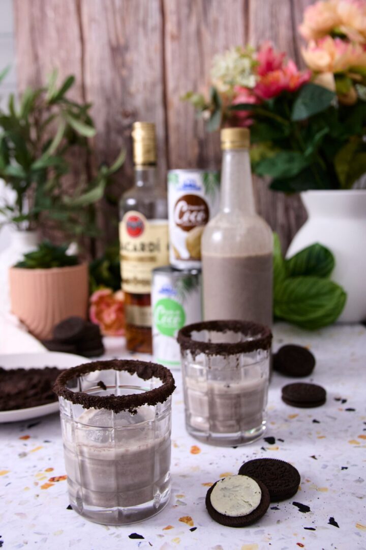 Two glasses rimmed with crushed cookies filled with cookies and cream coquito, with rum, coconut milk cans, and flowers blurred in the background.