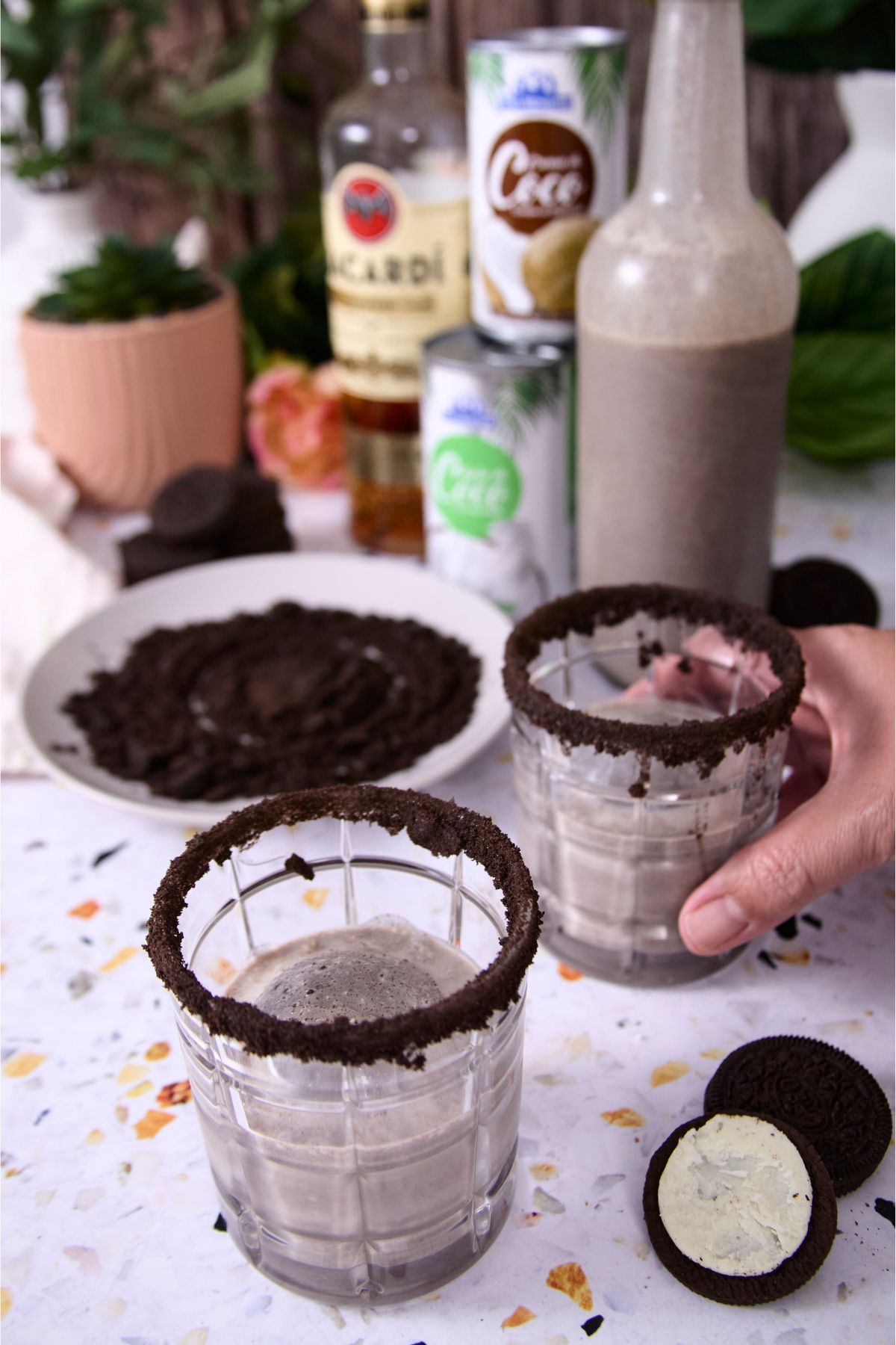 A hand picking up a glass of cookies and cream coquito with a cookie crumb rim, with crushed cookies on a plate, and ingredient cans behind it.