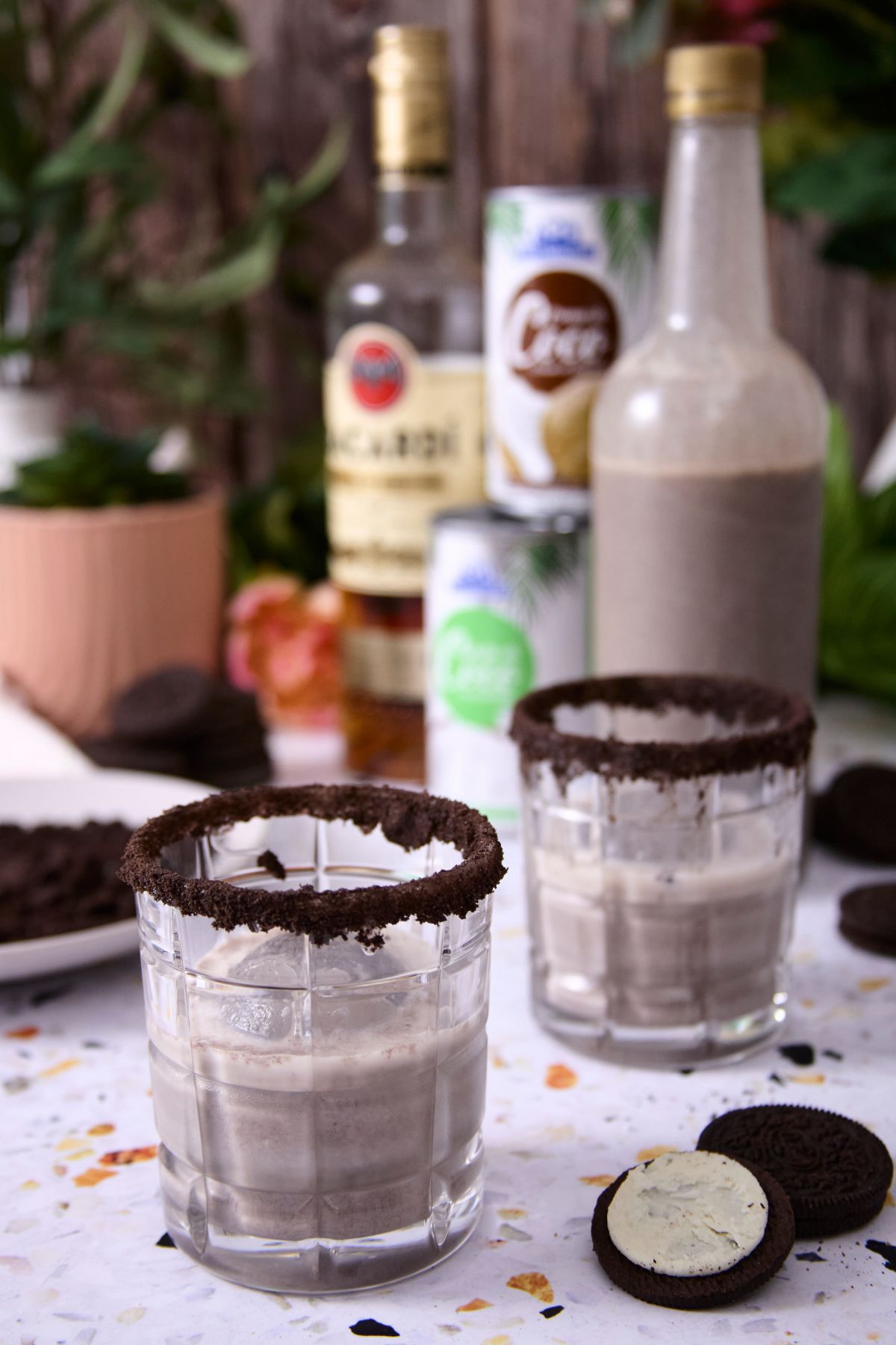 Two glasses of cookies and cream coquito with cookie crumb rims, surrounded by Oreo cookies and coquito ingredients in the background.