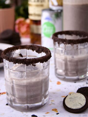 Two glasses of cookies-and-cream coquito with crushed-cookie rims, surrounded by Oreo cookies and coquito ingredients, blurred in the background.