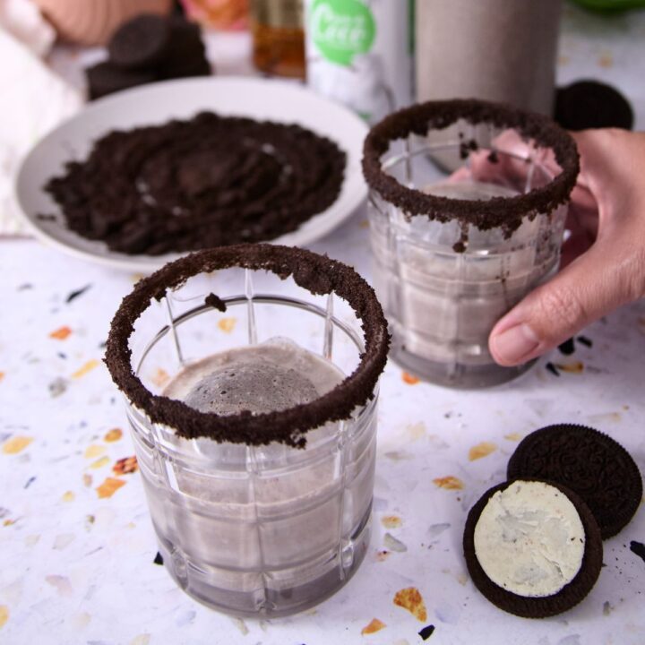 A hand lifting a glass of cookies-and-cream coquito with a crushed-cookie rim, with a plate of crushed cookies and Oreo halves nearby.