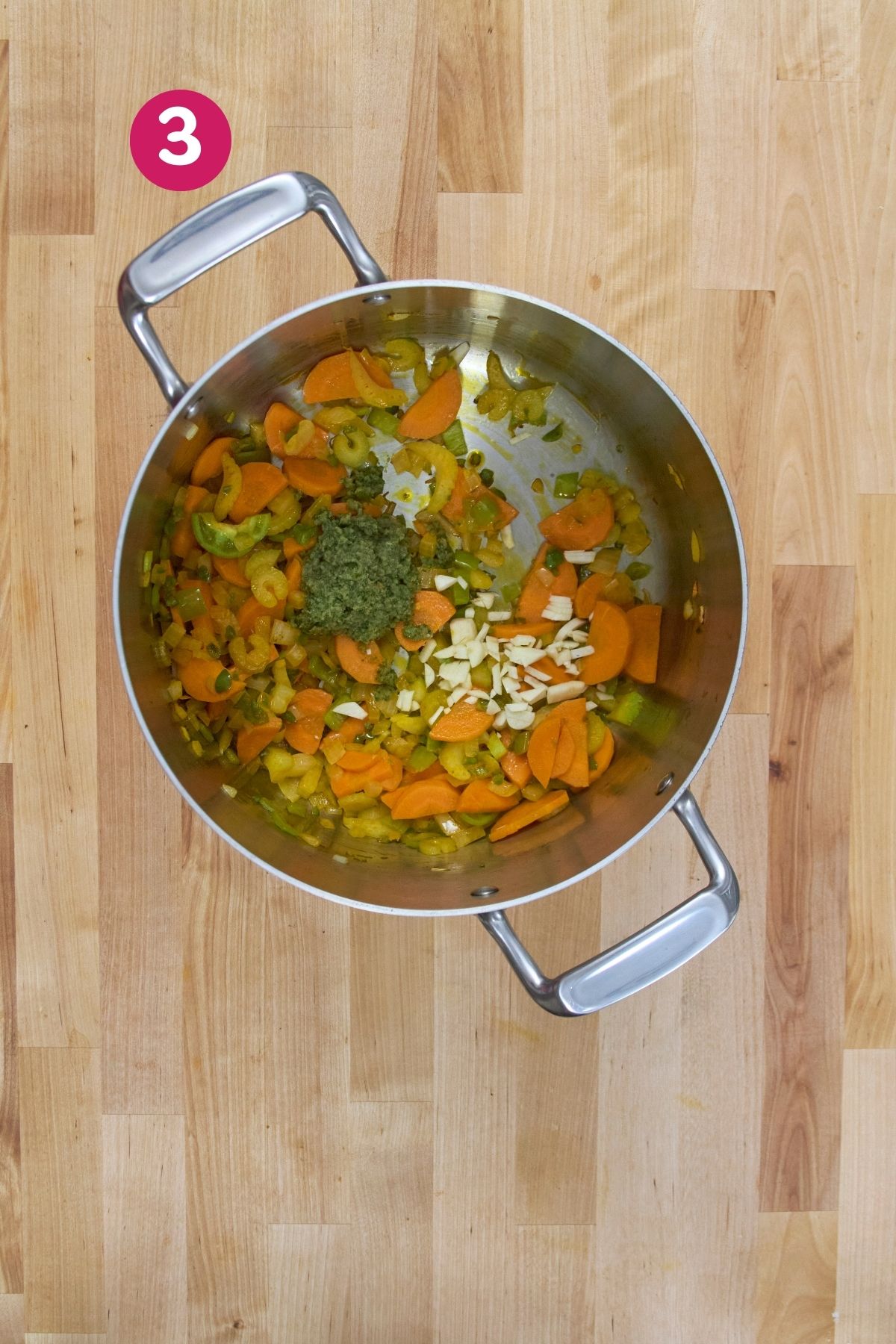 Pot with sautéed vegetables, chopped garlic, and sofrito added, viewed from overhead on a wooden countertop.
