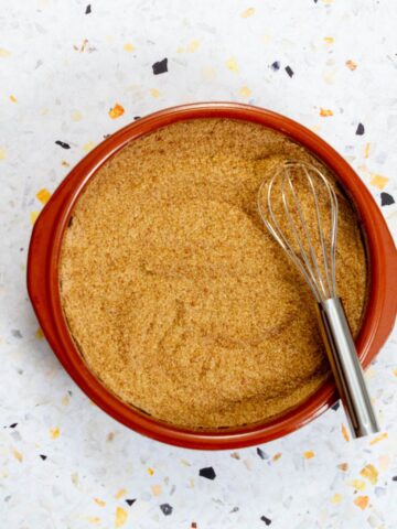 Top-down view of a shallow brown bowl filled with a golden, blended seasoning, with a metal whisk resting inside, on a light terrazzo background.