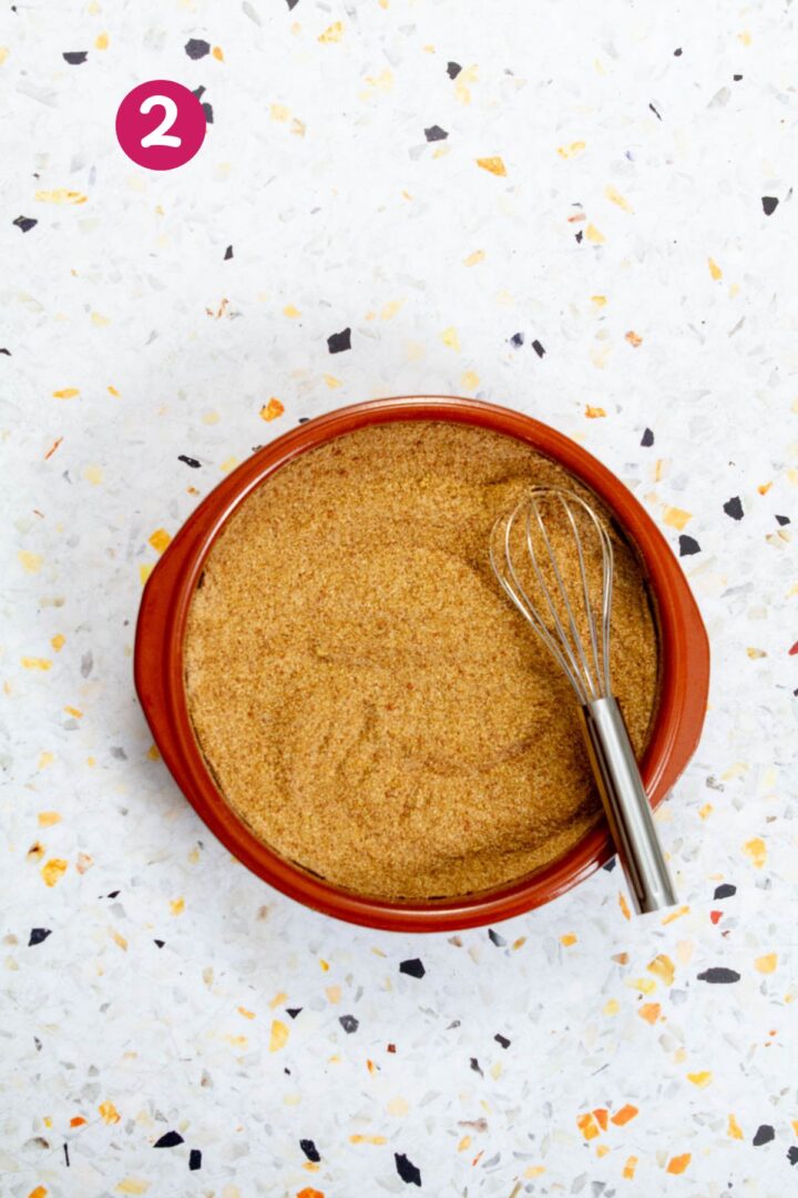 Top-down view of a shallow brown bowl filled with a golden, blended seasoning, with a metal whisk resting inside, on a light terrazzo background.