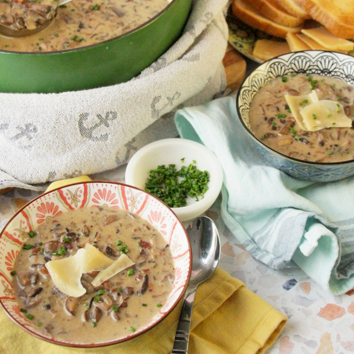 Styled overhead scene of bacon mushroom soup in a Dutch oven with a ladle, two bowls of soup garnished with shaved cheese and chives, bread slices, and folded kitchen towels.