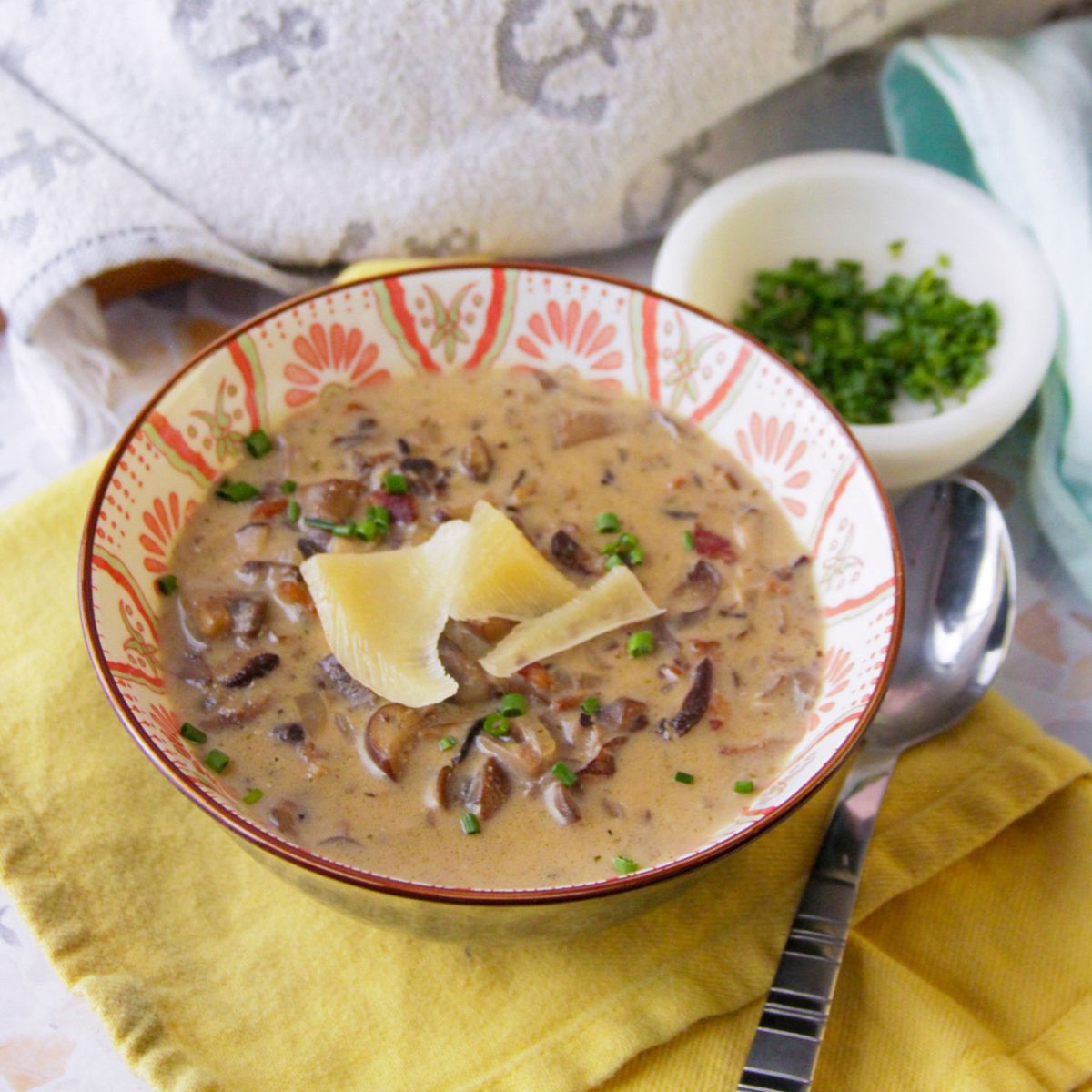 Overhead composition showing a green Dutch oven of bacon mushroom soup, two garnished bowls of soup, sliced bread, cheese, and a small bowl of chopped herbs on a countertop.
