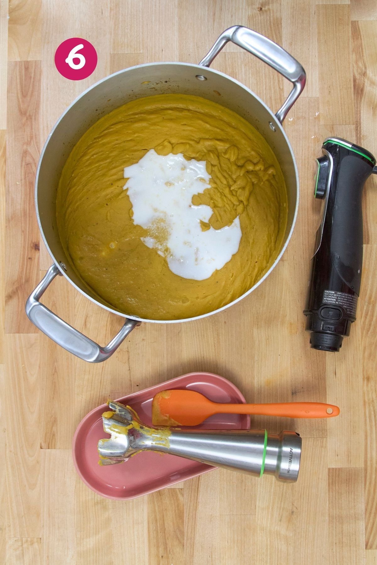 Pot of blended plantain soup with coconut milk added, immersion blender resting nearby on a wooden surface.