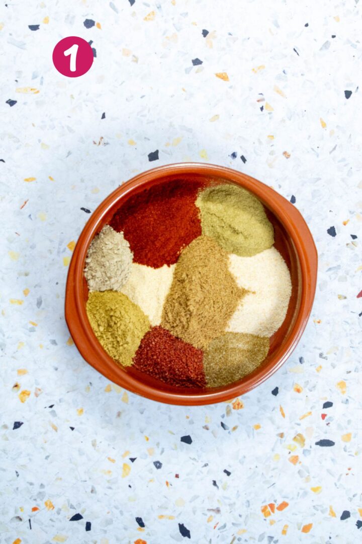 Top-down view of a brown bowl containing separate piles of ground spices arranged in sections, including red, beige, and green powders, on a light terrazzo surface.