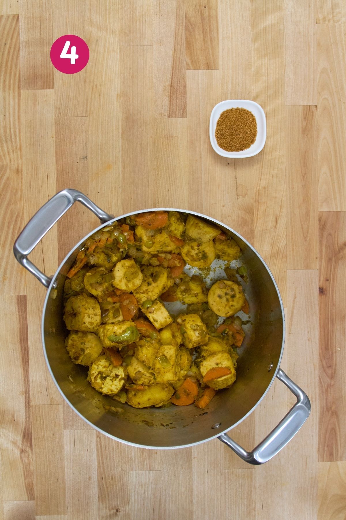 Pot with cooked plantain chunks and vegetables, with a small bowl of seasoning placed above on a wooden surface.