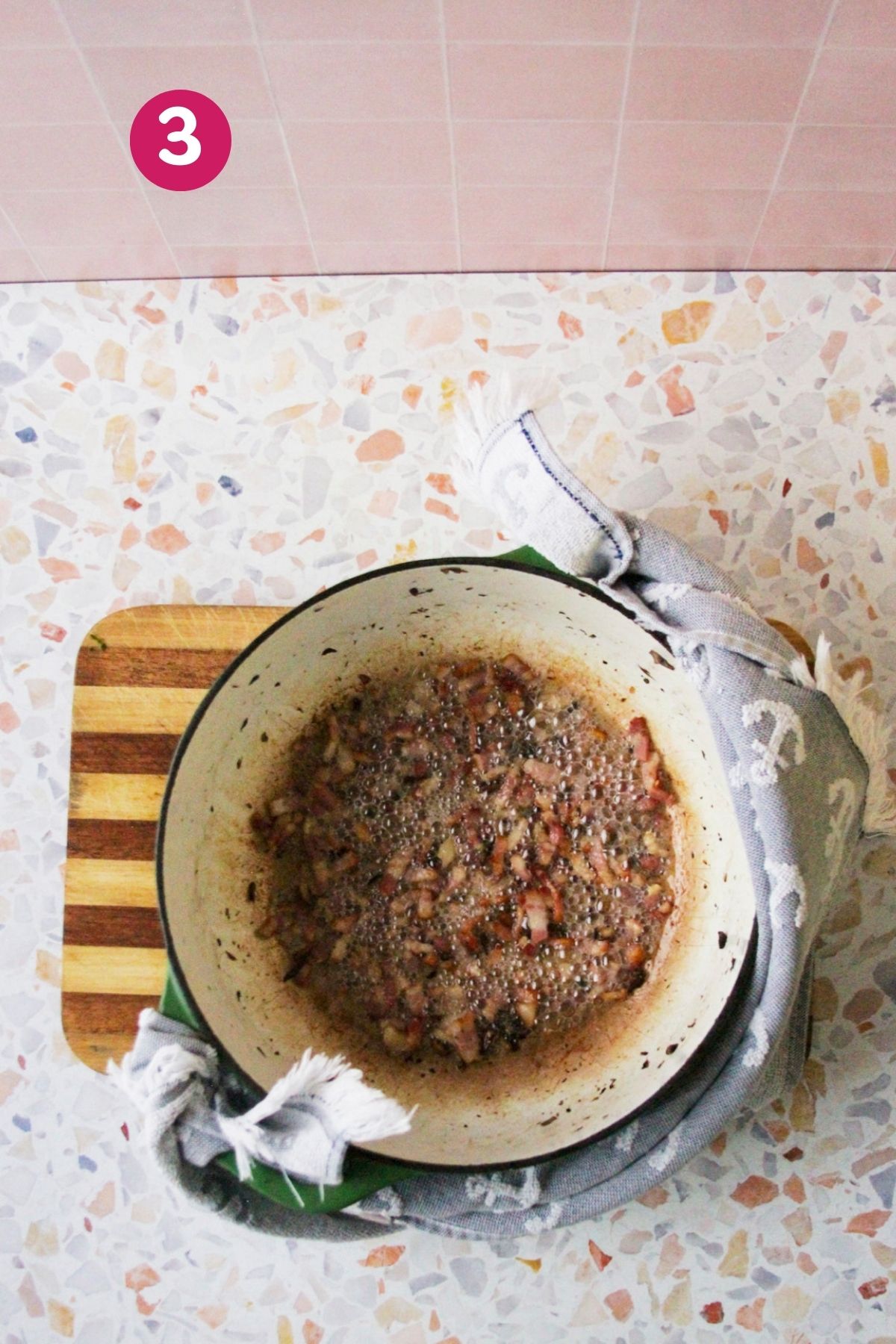 Bacon pieces rendering and bubbling in a Dutch oven on a cutting board, photographed from overhead.