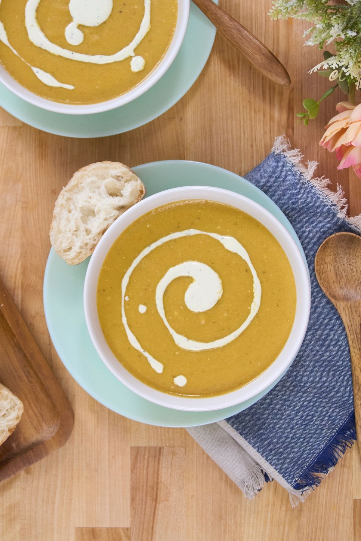 Two bowls of blended plantain soup topped with a cream swirl, served on light blue plates with bread on a wooden table.