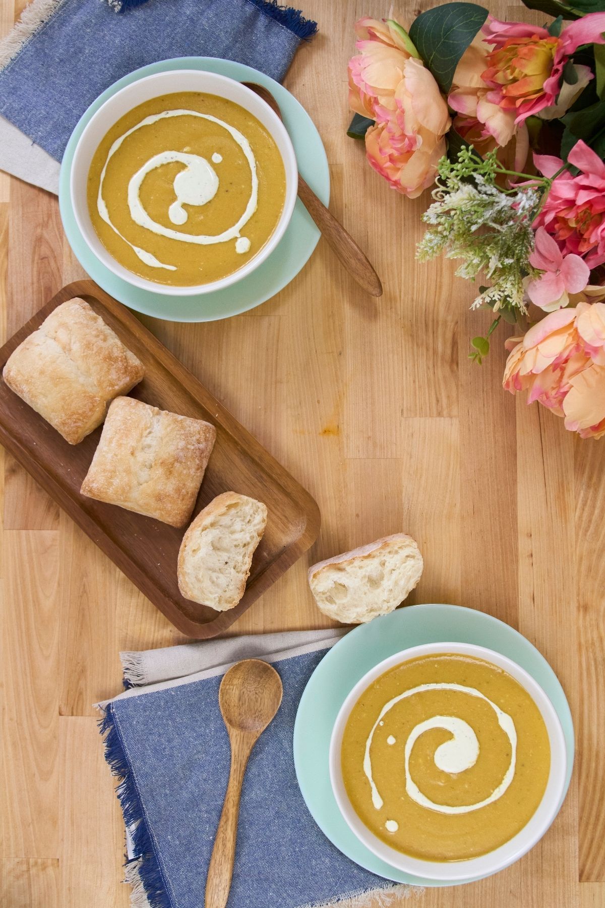 Overhead view of two bowls of plantain soup with cream swirl, bread pieces, wooden spoon, and folded blue napkins on a wooden surface.