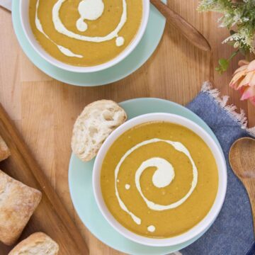 Overhead view of two bowls of plantain soup with cream swirl, bread pieces, wooden spoon, blue napkins, and flowers on a wooden table.