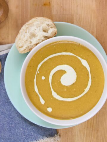 Overhead view of a single bowl of plantain soup with a cream swirl, bread slice on the rim, and wooden spoon on a blue napkin.