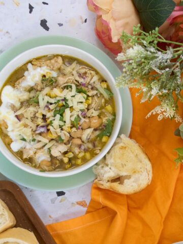 Overhead view of two bowls of white bean chicken chili served on green plates with bread slices on a cutting board, and flowers arranged on an orange cloth.