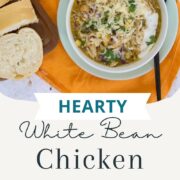 Bowl of white bean chicken chili topped with shredded cheese, chopped cilantro, diced red onion, and sour cream on an orange cloth with sliced bread on a wooden board.