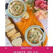 Two bowls of white bean chicken chili on a tabletop with sliced bread on a cutting board, an orange fabric napkin, and pink and peach flowers arranged to the side.