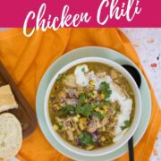 Close-up overhead view of white bean chicken chili garnished with cilantro, corn, red onion, shredded cheese, and sour cream in a light green bowl on an orange cloth.