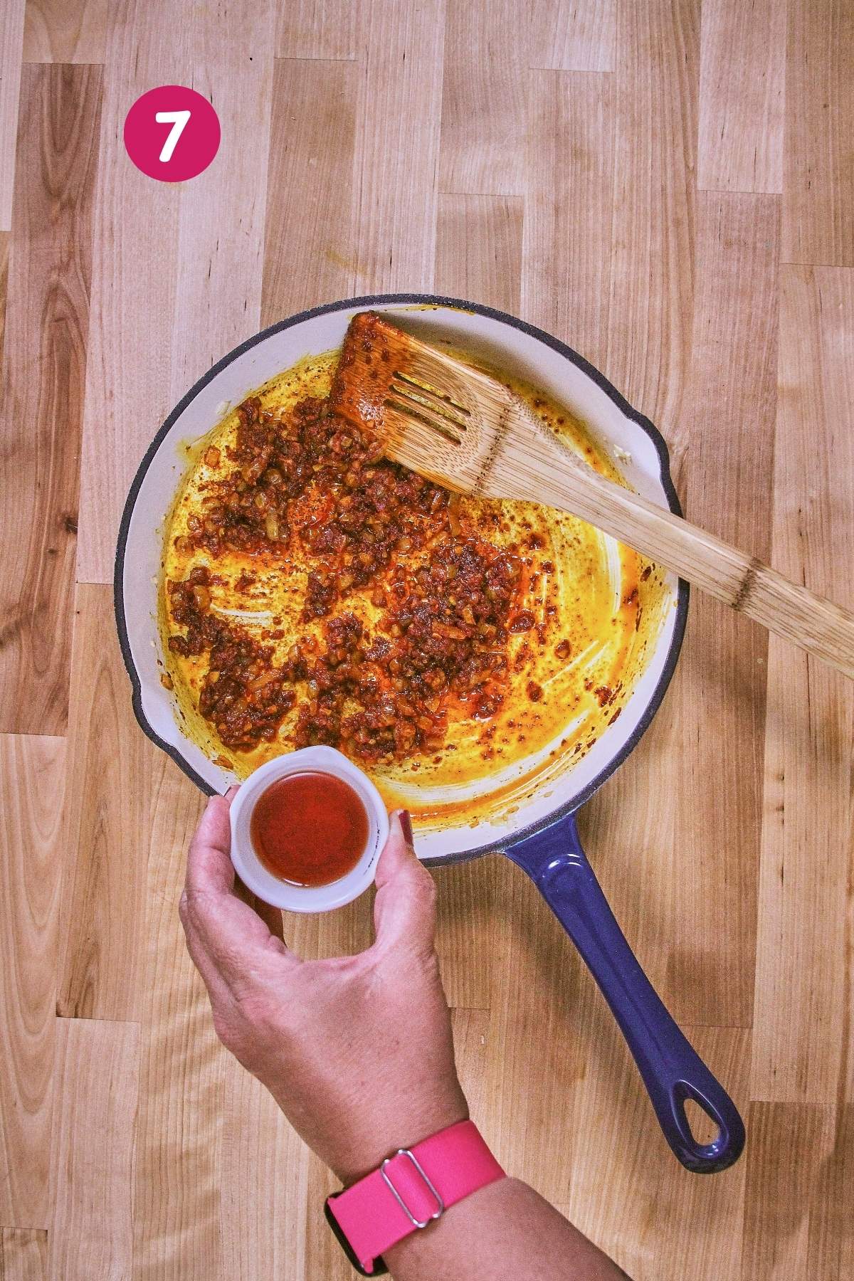 Hand holding a small cup of orange achiote oil over a skillet with sautéed shallots and tomato paste for brava sauce, labeled with step 7.