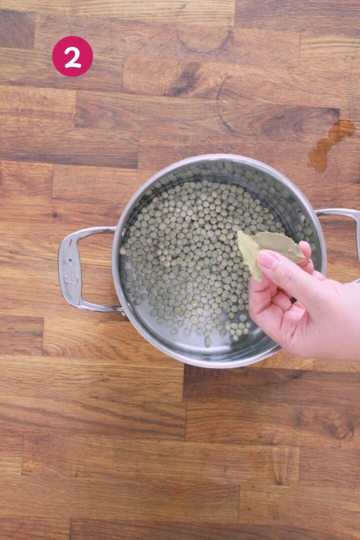 Hand adding bay leaves to pot of dried gandules (pigeon peas) in water for quick soak method, labeled with step 2.