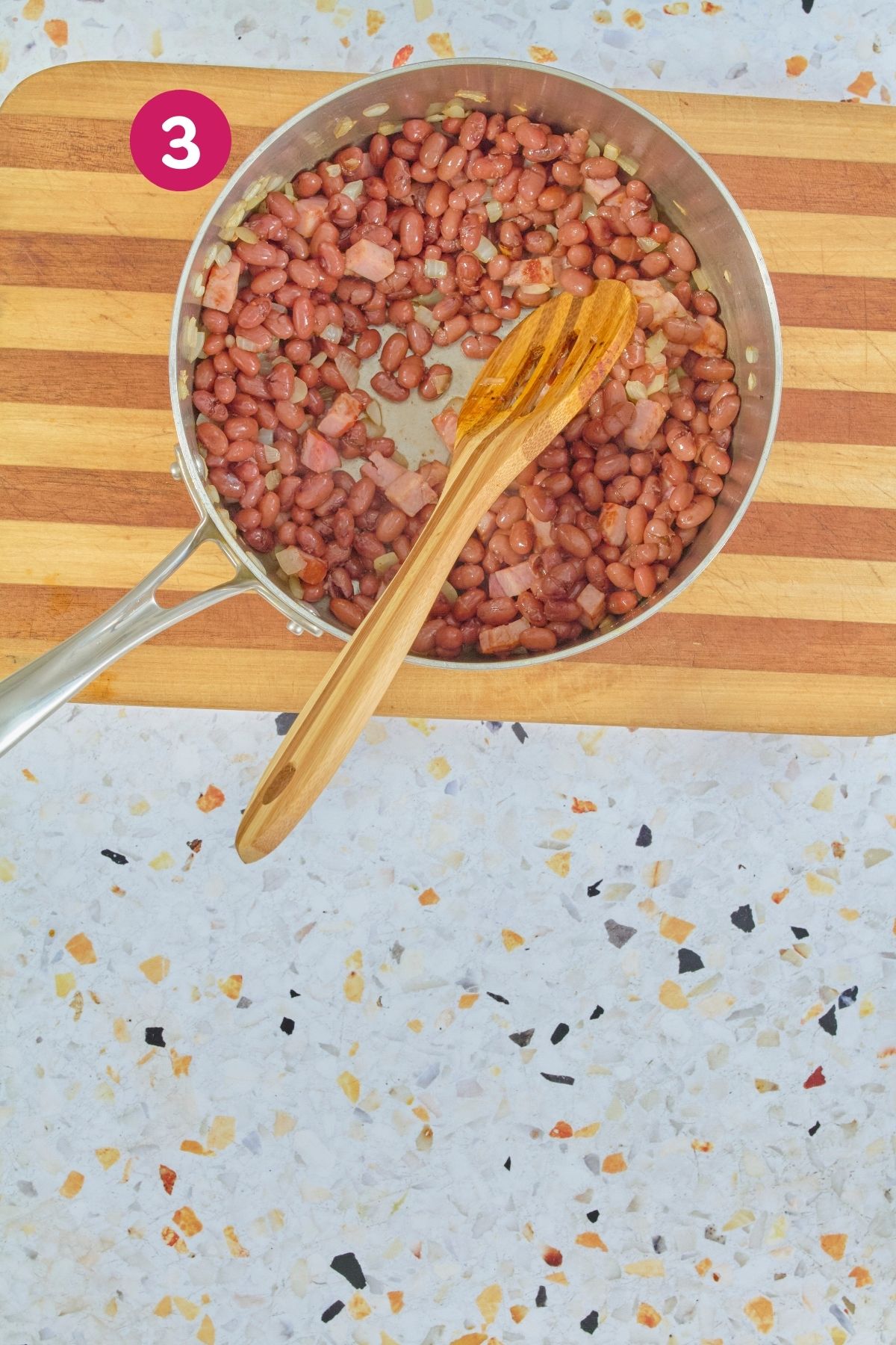 Overhead view of pink beans being added to sautéed onions and diced ham in a stainless steel pot, with a wooden spoon, labeled "step 3".