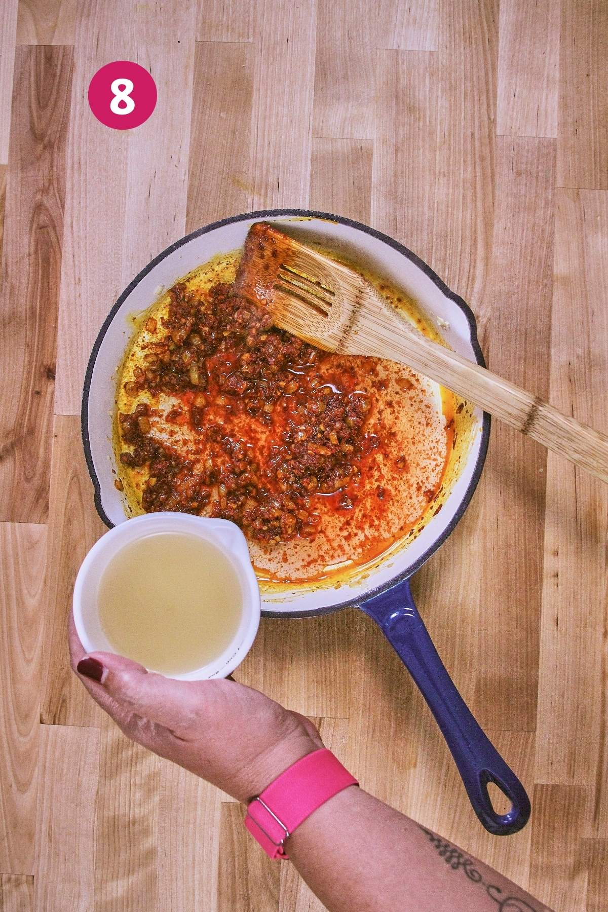 Hand holding white cup of broth over skillet with sautéed shallots, tomato paste and spices for brava sauce, labeled with step 8.