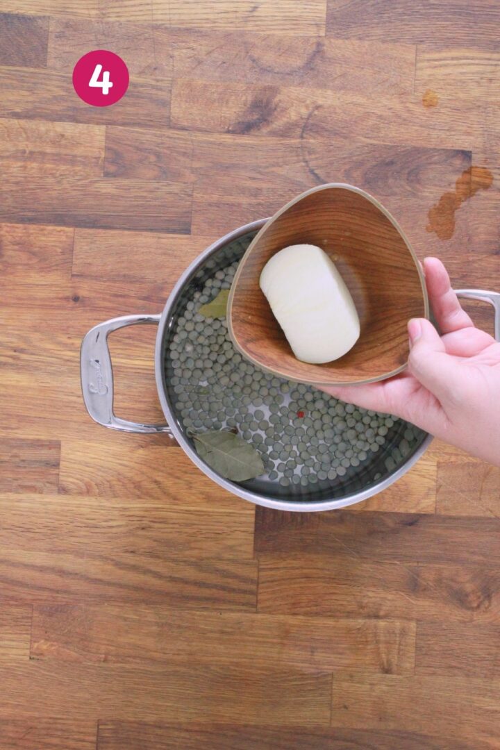 Hand holding wooden bowl with peeled whole onion over pot of soaking gandules with bay leaves and peppercorns, labeled with step 4.