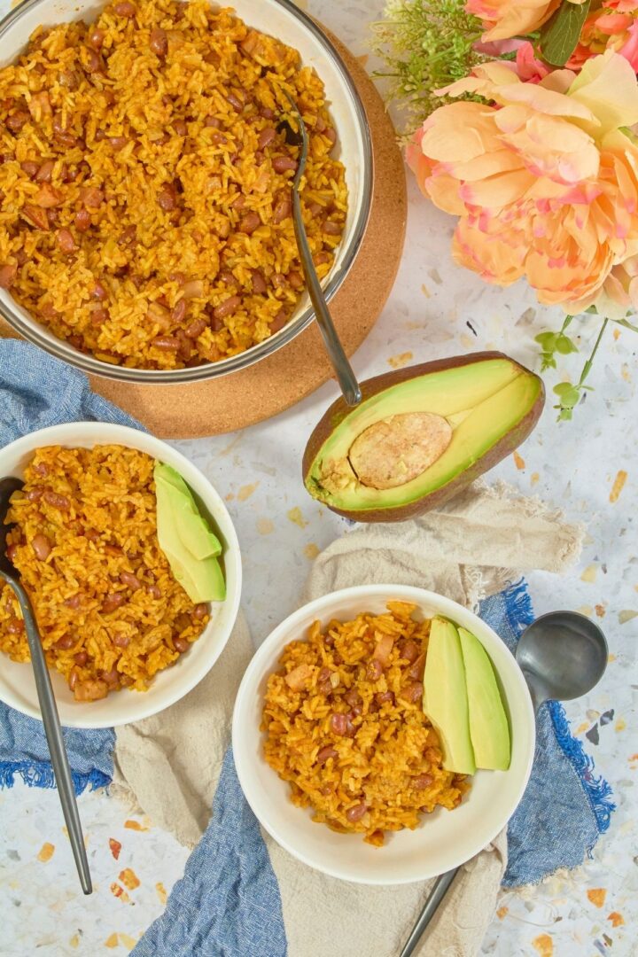 Overhead view of finished arroz con habichuelas in glass bowl and white bowls with avocado slices, avocado half and peach peonies on side.