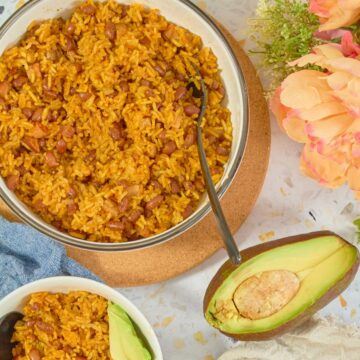 Close-up of arroz con habichuelas in a ceramic bowl with serving spoon, avocado half, and individual serving with avocado slices visible.