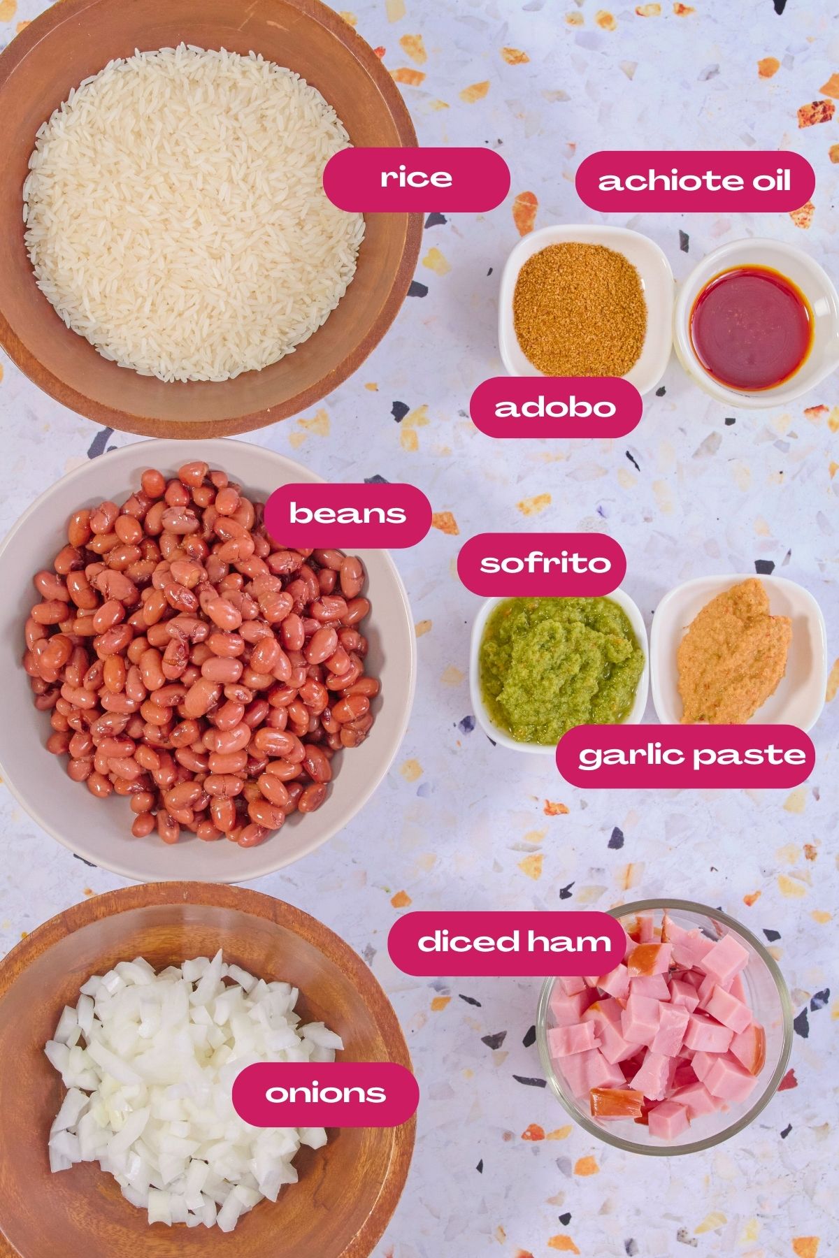 Overhead view of ingredients for arroz con habichuelas including white rice, pink beans, diced ham, onions, sofrito, garlic paste, adobo seasoning, and achiote oil in separate bowls.