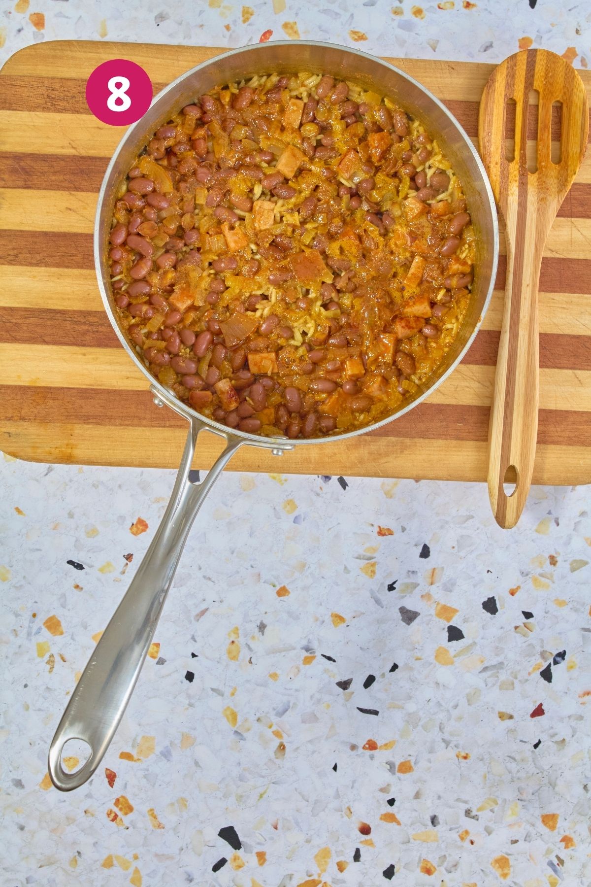 Overhead view of rice and beans mixture cooking in stainless steel skillet with excess moisture drying before covering, labeled with step 8.