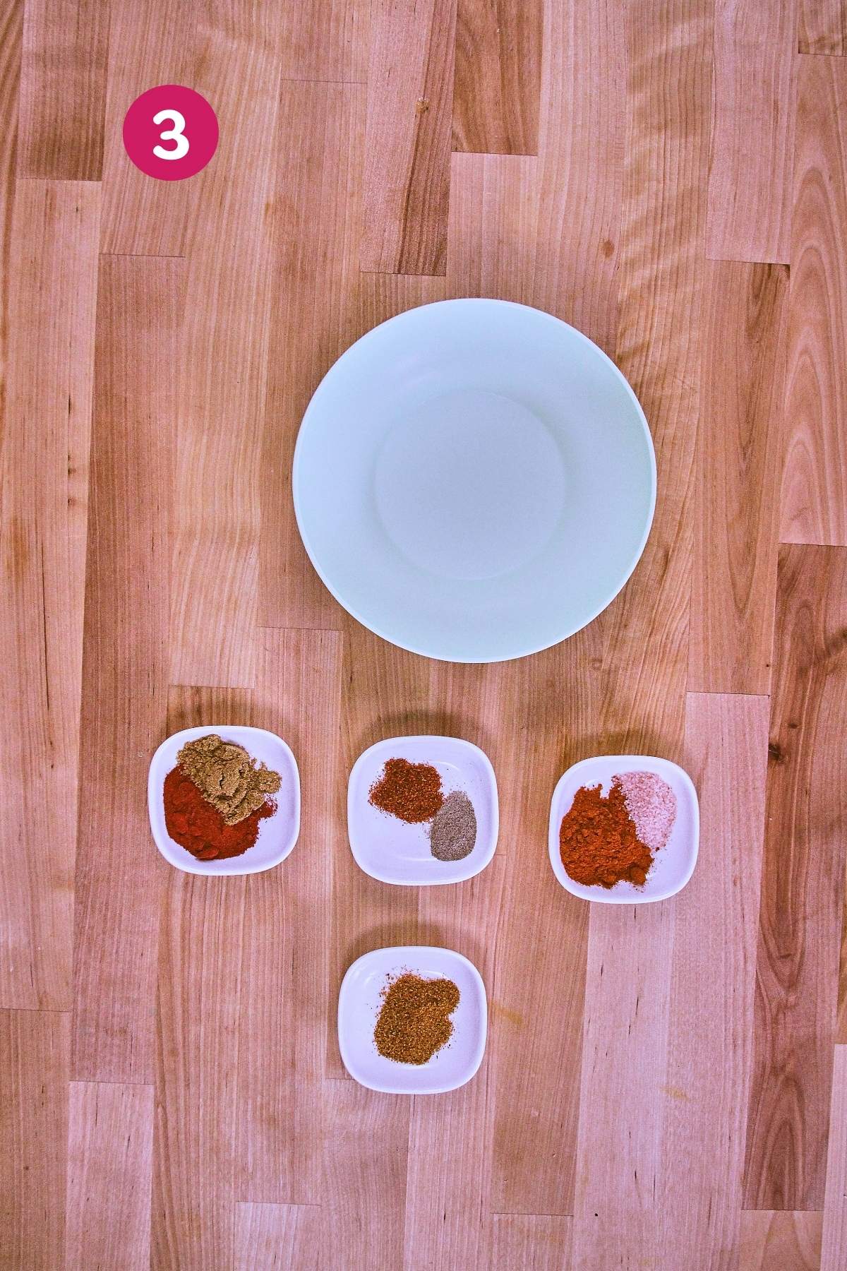 Overhead view of empty white plate with four small bowls of spices including smoked paprika, cumin, garlic powder and cayenne pepper arranged below, labeled with step 3.