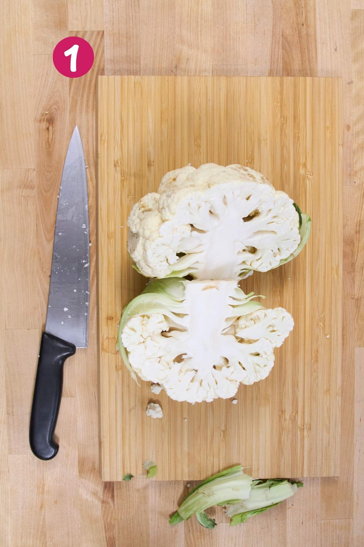 Overhead view of cauliflower head cut in half on bamboo cutting board with chef's knife and separated stem pieces, labeled with step 1.