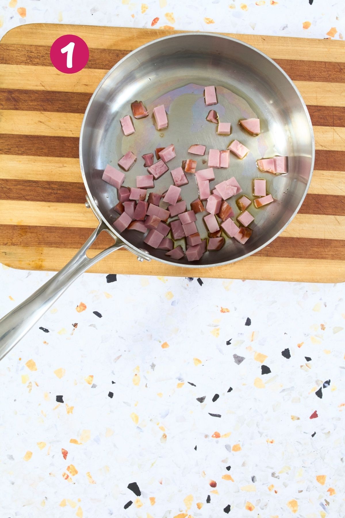 Overhead view of diced ham browning in stainless steel skillet with oil, labeled with step 1.