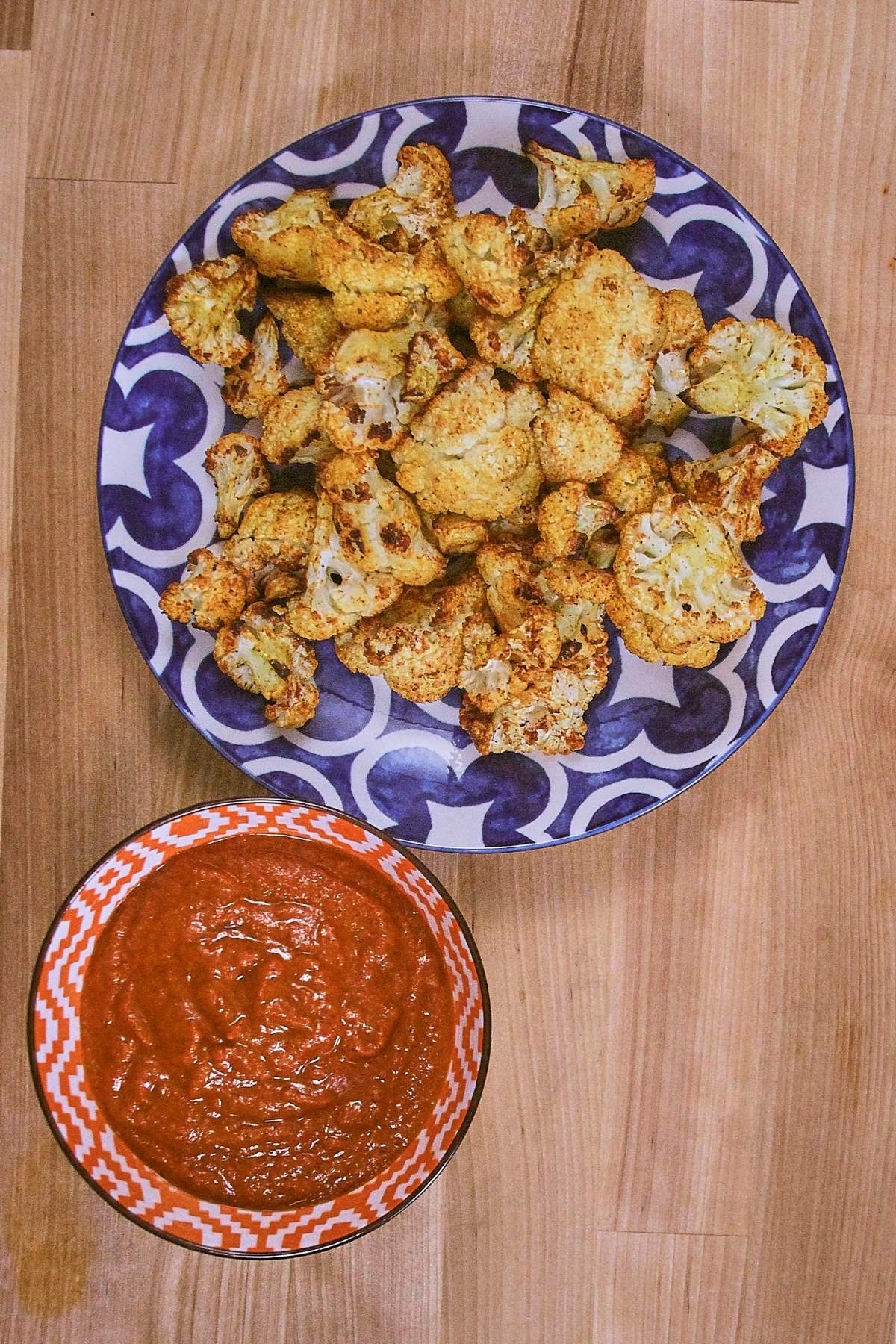 Overhead view of crispy roasted cauliflower florets on blue patterned plate with spicy brava sauce in an orange checkered bowl.